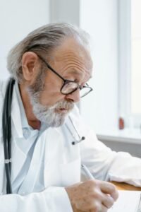 Senior doctor with eyeglasses writing notes during consultation indoors.